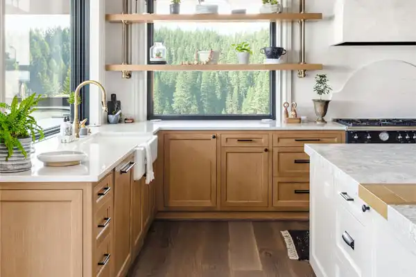 Modern kitchen with wooden cabinets, white countertops, a farmhouse sink, and a stove. Open shelves display plants and kitchenware. Large window offers a view of trees outside.